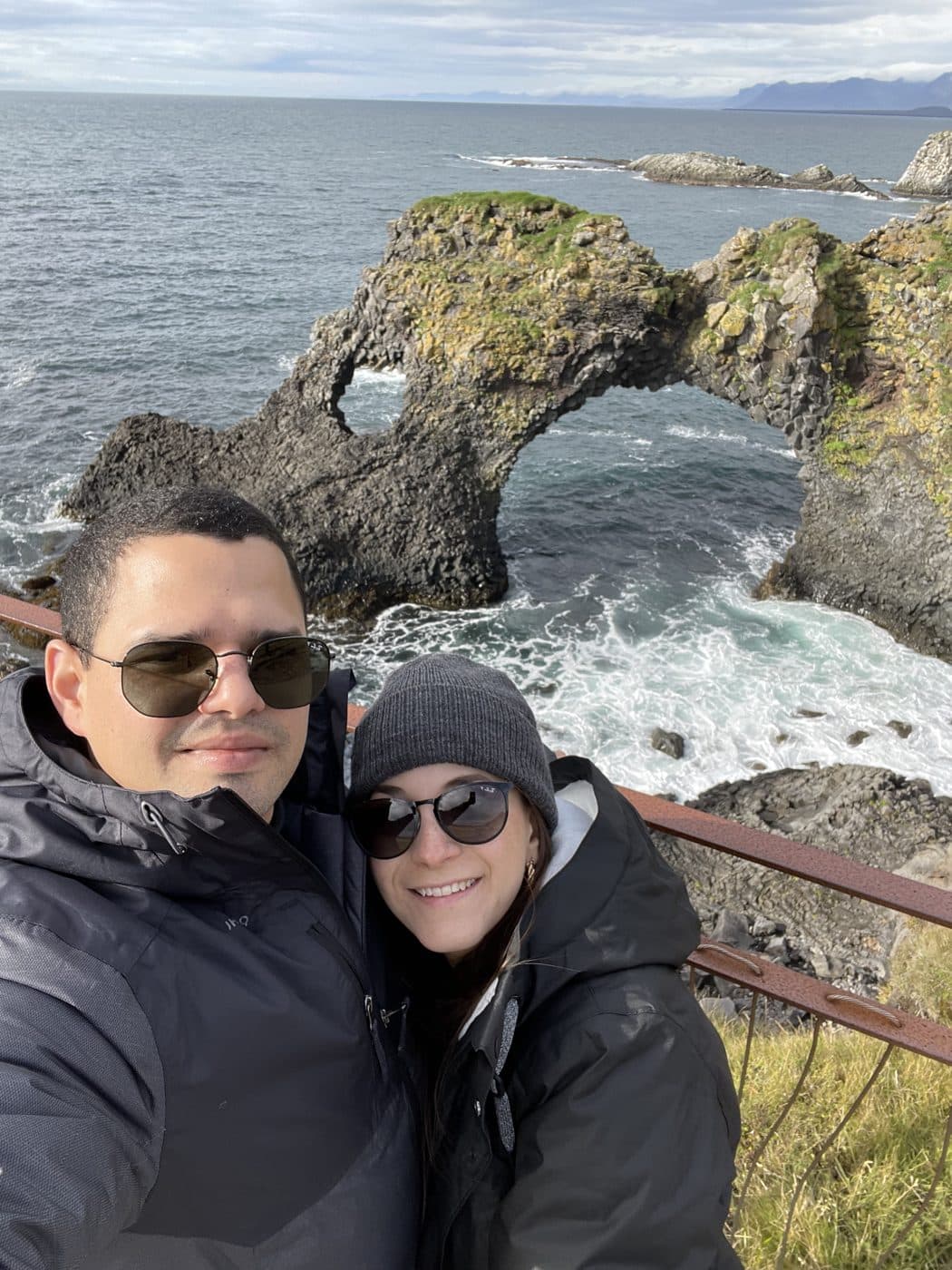 Adriana and Yair in winter coats at a railing above a sea arch.