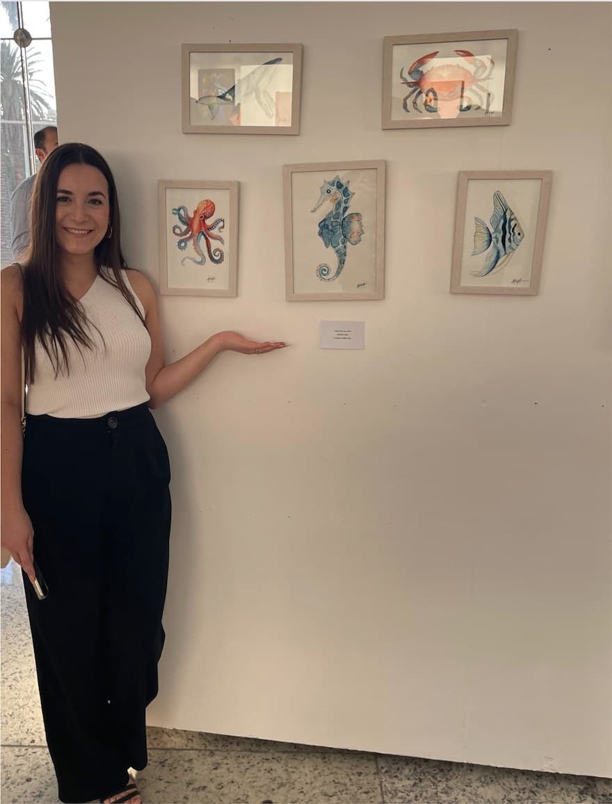 Adriana beside a wall of her framed watercolours at an exhibition.