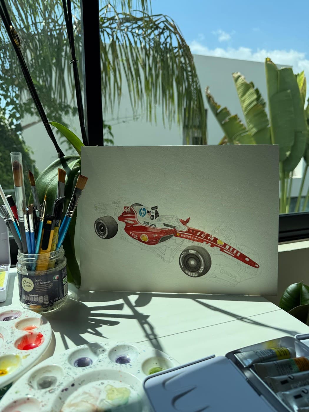 A half-finished watercolour of a red Formula 1 car propped on a desk, surrounded by brushes and paint palettes, with sunlit palms through the window behind it.